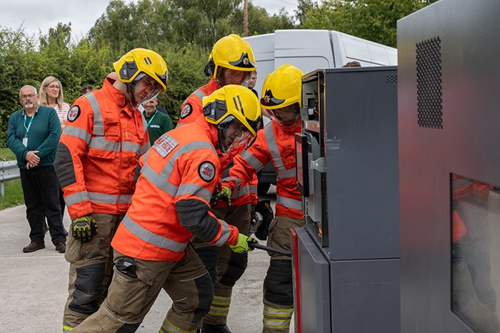 Firefighters in protective gear attempt to force open an ATM unit using tools during a live demonstration, while observers watch nearby.