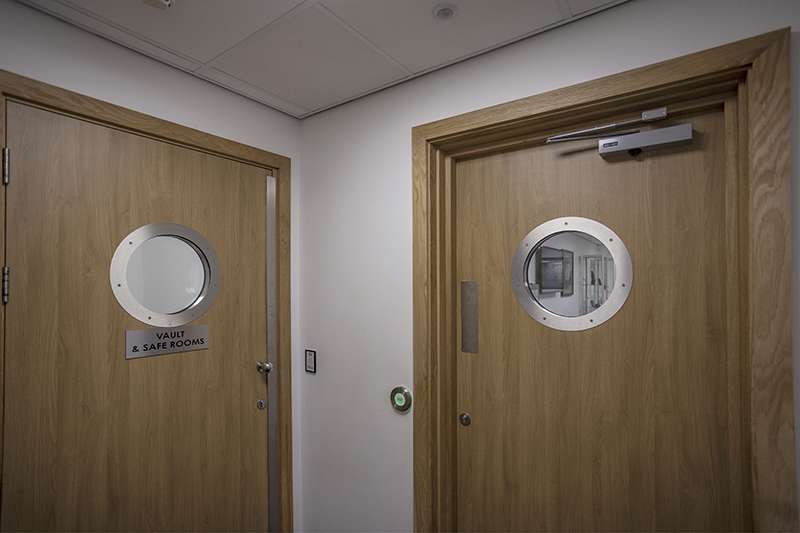 Two wooden security doors with circular reinforced windows in a secure facility, one labeled Vault & Safe Rooms.